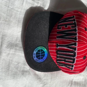 Röd och svart keps med New York-tryck - Snygg röd keps med vita ränder och stort New York-tryck framtill. Har aldrig kommit till användning!