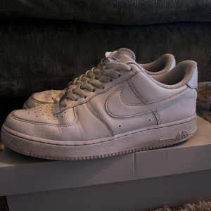 Vita Nike Air Force 1 '07 - Säljer ett par klassiska vita Nike Air Force 1 '07. Skorna har en stilren design med snörning och perforerade detaljer på tån för ventilation. Perfekta för en casual look. Man får med ett par skydd till framsidan av skon också