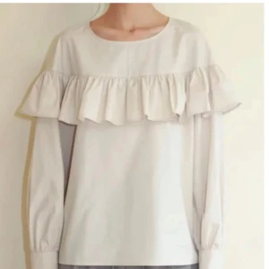 Vit blus med volangdetalj - Säljer en stilren vit blus med volangdetalj över bröstet och långa ärmar. Perfekt för en elegant look. Blusen har en rund halsringning.