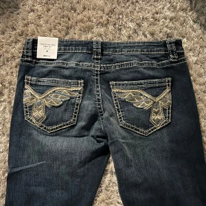 Lowrise jeans med broderade bakfickor - Snygga lowrise mörkblå jeans med unika broderade detaljer på bakfickorna. Byxorna har en bootcut-stil som ger en klassisk look. Perfekta för dig som vill ha något extra i garderoben. Midjemått ca 39cm och Innerbenslängd ca 78-80cm.