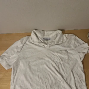 Vit frotté tröja - Tja! säljer en as bekväm vit t-shirt från ST Studios i storlek L. T-shirten har korta ärmar och en klassisk krage med en bröstficka. Tröjan är gjord av frotté på utsidan och bomull på insidan. Perfekt för en sval sommarkväll.