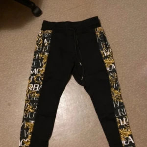 Svarta mjukisbyxor från Versace Jeans Couture - Snygga svarta mjukisbyxor från Versace Jeans Couture med guld- och vitmönstrade sidopaneler. Byxorna har en bekväm midwaist och snörning i midjan för justerbar passform. Perfekta för en stilren och avslappnad look.