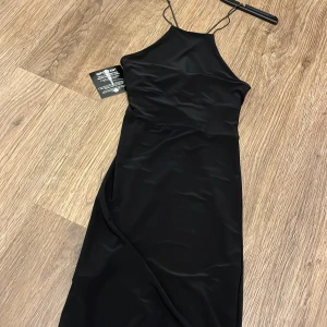 Svart oneshoulder klänning - Elegant svart oneshoulder klänning med smala axelband. Perfekt för en stilren look. Klänningen har en slät yta och är figurnära.