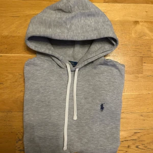 Grå hoodie från Ralph Lauren - Säljer en stilren grå hoodie från Ralph Lauren med den klassiska broderade loggan på bröstet. Tröjan har en justerbar huva med snören.