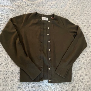 Olivgrön kofta från Holly & Whyte - Säljer en stilren olivgrön kofta från Holebrook. Koftan har långa ärmar och knappar framtill. Perfekt för lager-på-lager och passar till många olika outfits. Ett måste i garderoben för den som gillar klassisk stil. Mycket fint skick, skulle nog säga använd 1-2 gånger!