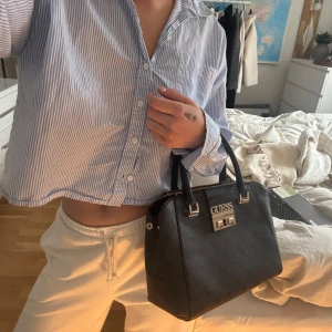 Svart handväska från Guess - Snygg svart handväska från Guess med elegant logotypdetalj framtill. Väskan har dubbla handtag och en praktisk dragkedja upptill. Insidan är rymlig med flera fack för enkel organisering. Perfekt för att bära med sig det nödvändigaste med stil. Lite skavanker på insidan**