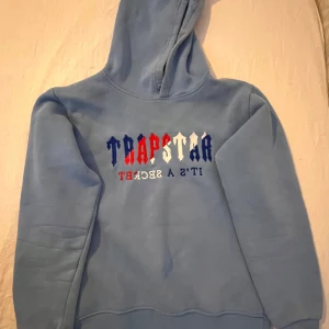 Blå hoodie från Trapstar - Säljer en blå hoodie från Trapstar med texten 'TRAPSTAR IT'S A SECRET' i rött, vitt och blått på ryggen. Hoodien har en klassisk design med huva och långa ärmar. Perfekt för en avslappnad stil.