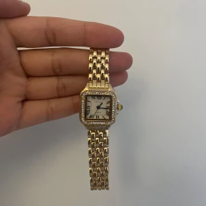 Guldfärgad klocka med glittrig ram - Elegant guldfärgad klocka med fyrkantig urtavla och romerska siffror. Klockan har en glittrig ram och en länkarmband i guld. Storlek går att ändra. Pris kan diskuteras vid snabb affär