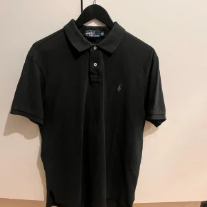 Svart pikétröja från Ralph Lauren - Säljer en klassisk svart pikétröja från Ralph Lauren med korta ärmar och en liten lila logga på bröstet. Tröjan har en stilren krage och knappar framtill. Perfekt för en avslappnad men ändå stilren look.