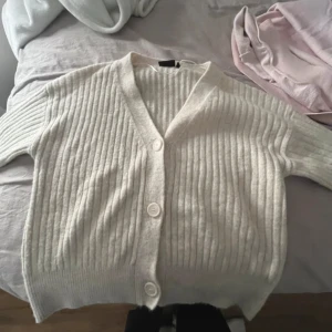 Vit ribbad kofta från KappAhl - Mysig vit kofta från KappAhl i storlek S. Den är ribbad med stora knappar framtill och har en v-ringad design. Perfekt för kyliga dagar när du vill hålla dig varm och stilren.