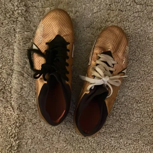 Guldiga mercurial mbappe fotbollskor från Nike - Snygga guldiga fotbollsskor från Nike med svart och vit detaljering. Skorna har snörning och en slitstark sula med bra grepp för spel på gräs. Perfekta för fotbollsspelare som vill sticka ut på planen.