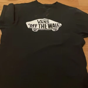 Svart t-shirt från Vans - Säljer en svart t-shirt från Vans med det klassiska 'Off The Wall' trycket i vitt på framsidan. T-shirten är tillverkad i 100% bomull och har en normal passform. Perfekt för en avslappnad stil.