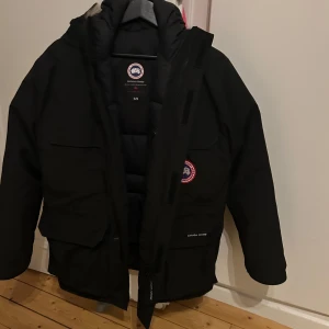 Svart dunjacka från Canada Goose - Säljer en svart dunjacka från Canada Goose med deras ikoniska logga på bröstet. Jackan har en långärmad design och är perfekt för kalla vinterdagar. Den har flera praktiska fickor och en dragkedja framtill. Är lite säker på storleken den är pojke xl sitter bra på båda dam och herr 