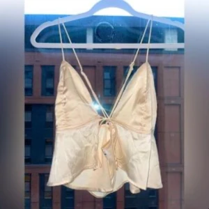 Vit satinlinnetopp med knyt - Säljer en vit satinlinnetopp med smala axelband och knytning framtill. Toppen har en djup v-ringning och en lätt glansig finish som ger en lyxig känsla. 