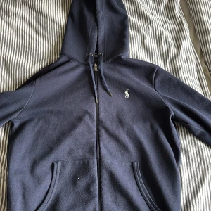 Mörkblå hoodie från Ralph Lauren - Säljer en stilren mörkblå hoodie från Ralph Lauren med dragkedja och huva. Den har en liten broderad logga på bröstet och praktiska fickor framtill. Perfekt för en avslappnad look. Nypris: 1500-1700, Pris kan diskuteras. Det är storlek M men funkar bra till S