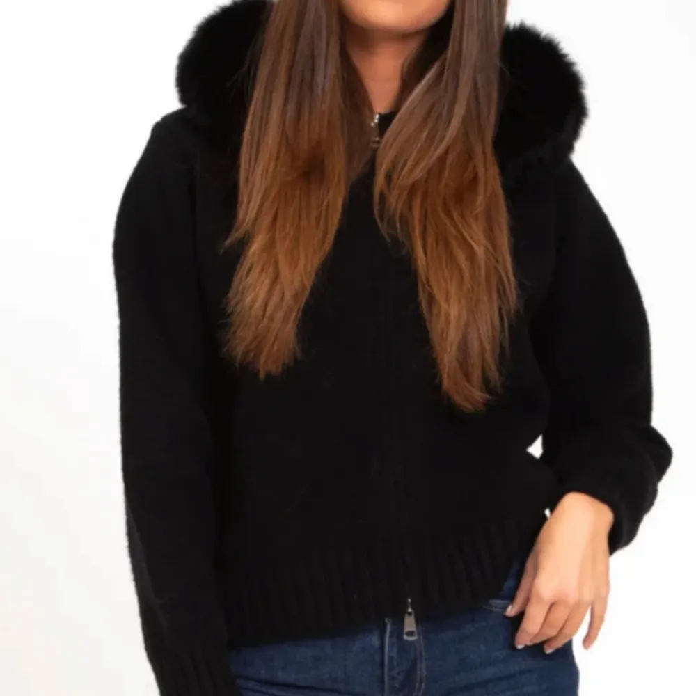 Snygg svart stickad jacka med en fluffig pälshuva. Jackan har dragkedja framtill och långa ärmar. Perfekt för kyliga dagar när du vill hålla dig varm och stilren. Jackan är köpt för 1800 kr och använd 2 gånger . Takit.