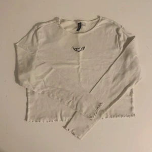 Vit långärmad topp - Säljer en vit långärmad topp från H&M med ett broderat hjärta med vingar. Toppen är croppad med vågiga kanter vid ärmslut och nederkant. Perfekt för en avslappnad stil! Storlek L men passar också M 