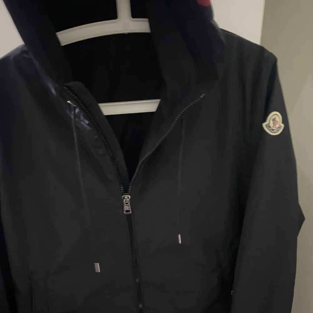 Säljer en stilren svart vindjacka från Moncler med dragkedja och huva. Jackan har Monclers logotyp på ärmen och detaljer i rött och vitt på huvan. Perfekt för blåsiga dagar.. Takit.