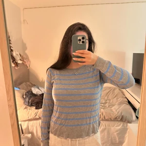 Grå och blå randig tröja - Säljer en stilren grå och blå randig tröja med långa ärmar. Perfekt för en avslappnad look. Tröjan har en enkel design med rund hals och är tillverkad i ett mjukt material.
