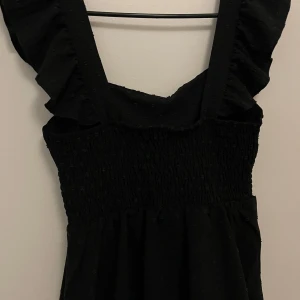 Svart topp från Skue - Snygg svart blus från Skue med volangdetaljer och smockad överdel.  Diskret prickigt mönster och är ärmlös med breda axelband. Perfekt för en stilren look. Storlek m men skulle säga att den är liten i storleken