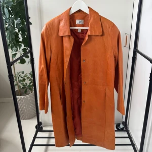 Orange skinnkappa  - Snygg orange skinnkappa med klassisk krage och knappar framtill. Perfekt för att ge en färgklick till din outfit. Perfekt nu till våren!