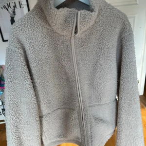 Brun teddyjacka med dragkedja - Mysig brun teddyjacka med dragkedja framtill. Perfekt för kyliga dagar med sin fluffiga och varma design. Jackan har en hög krage och långa ärmar för extra värme. Prislapp kvar!