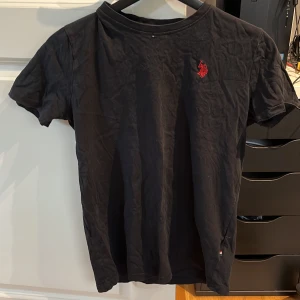 Svart kortärmad t-shirt polo - Snygg svart t-shirt med korta ärmar och en röd broderad polo logga på bröstet. Perfekt för en avslappnad stil. T-shirten har en klassisk rund halsringning.