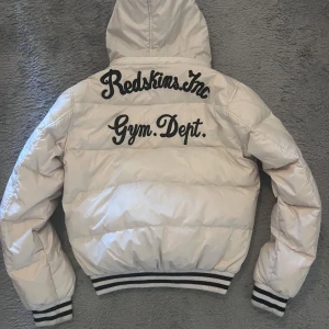 Vit pufferjacka från Redskins - Säljer en snygg vit pufferjacka från Redskins med text på ryggen och bröstet. Jackan har en huva och dragkedja framtill. Ribbstickade muddar vid ärmslut och nederkant ger en sportig look. Perfekt för kyliga dagar! Köptes för 1500kr 