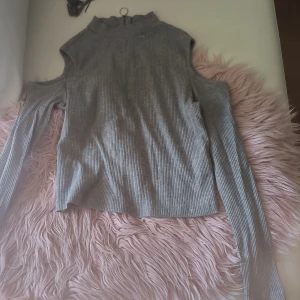 Grå ribbad topp från Divided - Säljer en grå ribbad topp från Divided med lång ärm och öppen axeldesign. Toppen har en dragkedja bak och en hög krage. Perfekt för en trendig look!