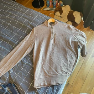 Brun tröja med dragkedja från Polo Ralph Lauren - Säljer en stilren brun tröja från Polo Ralph Lauren med halv dragkedja och broderad logga på bröstet. Perfekt för en avslappnad men snygg look. Tröjan har långa ärmar och är tillverkad i ett mjukt material.