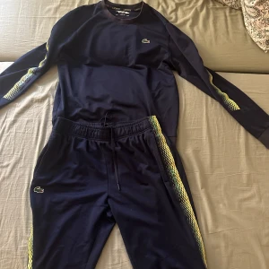 Mörkblå träningsset från Lacoste - Säljer ett mörkblått träningsset från Lacoste Sport med gula detaljer längs ärmar och byxben. Tröjan har långa ärmar och en klassisk passform. Perfekt för sportiga aktiviteter eller en avslappnad dag. Passar S och M tycker jag
