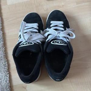 Svarta sneakers från Adidas - Snygga svarta sneakers från Adidas med vit sula och vita snören. Skorna har den klassiska Adidas-loggan på tungan och hälen. Perfekta för en avslappnad stil.