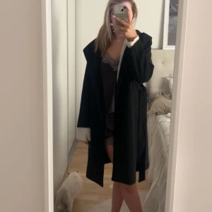 Svart kappa med bälte - Elegant svart kappa med bälte som ger en stilren look. Perfekt för kyligare dagar med sin långa design och klassiska snitt. Den har en enkel och tidlös stil som passar till många olika outfits.