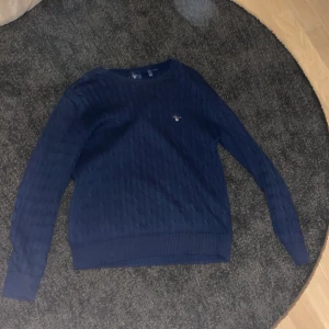 Mörkblå stickad tröja från GANT - Säljer en stilren mörkblå stickad tröja från GANT med kabelstickat mönster. Tröjan har långa ärmar och en broderad logotyp på bröstet. Perfekt för kyligare dagar.