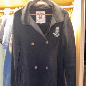 Svart cardigan från Morris - Säljer en stilren svart cardigan från Morris med dubbelknäppning och broderad detalj på bröstet. Jackan har långa ärmar och en klassisk krage. Perfekt för en elegant och avslappnad stil.