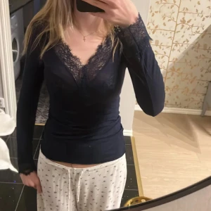 Mörkblå långärmad topp med spetsdetaljer - Säljer en elegant mörkblå topp med v-ringning och spetsdetaljer vid halsen och ärmsluten. Perfekt för en stilren look. Toppen har en figurnära passform och långa ärmar. Även väldigt skönt material! Original pris är 489kr 