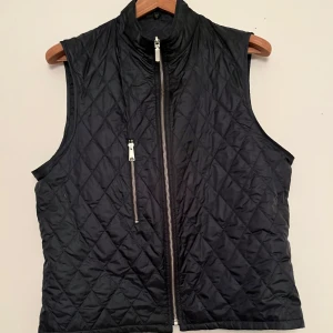 Svart quiltad väst med dragkedja - Snygg mörkblå quiltad väst med dragkedja framtill. Perfekt för lager-på-lager och ger en stilren look. Västen är tunn och är perfekt nu för våren. Västen har inga defekter och har knappt används