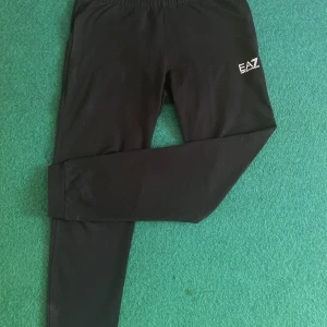 Svarta mjukisbyxor från Emporio Armani - Snygga svarta mjukisbyxor från Emporio Armani med EA7-logga på sidan. Byxorna har elastisk midja och är perfekta för en avslappnad stil. De har även ett diskret randigt mönster på baksidan. Storleken är S men passar även M!
