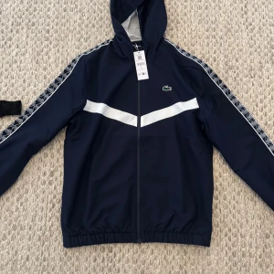 Blå vindjacka från Lacoste - Säljer en snygg blå vindjacka från Lacoste med vit rand över bröstet och längs ärmarna. Jackan har en huva och dragkedja framtill. Perfekt för en sportig look!
