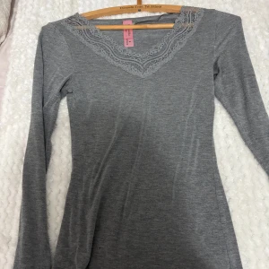 Grå långärmad topp från Hunkemöller - Säljer en stilren grå långärmad topp från Hunkemöller med vacker spetsdetalj vid halsringningen. Perfekt för en elegant och bekväm look. Passar bra till både jeans och kjol. Endast använd 1 gång 