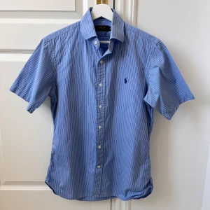 Blå randig skjorta från Polo Ralph Lauren - Riktigt ny skick! Snygg blå kortärmad skjorta från Polo Ralph Lauren med vita ränder och klassisk krage. Skjortan har knappar framtill och en broderad logotyp på bröstet. Perfekt för en stilren look! Rekommenderar för M 