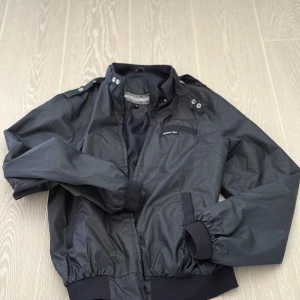 Svart Members only jacket sopranos - Snygg svart bomberjacka från Members Only med dragkedja och knappar. Jackan har långa ärmar och ribbade muddar vid ärmslut och nederkant. Perfekt för en stilren look.