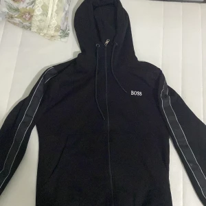 Svart zip hoodie från Hugo Boss - Snygg svart hoodie från Boss med dragkedja och huva. Den har vita detaljer längs ärmarna och en broderad logga på bröstet. Perfekt för en stilren och bekväm look.Och har använt den typ 4 gånger bara