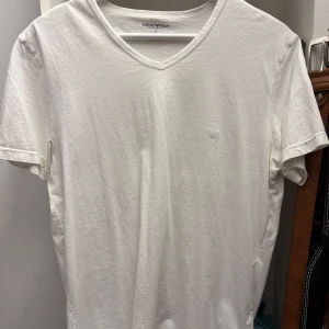 Vit v-ringad t-shirt från Emporio Armani - Säljer en stilren vit t-shirt från Emporio Armani med v-ringning. T-shirten är tillverkad i mjuk bomull och har en diskret logga på bröstet. Perfekt för en minimalistisk look.