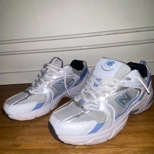 Vita och blå sneakers från New Balance - Snygga vita sneakers från New Balance med blå detaljer och klassisk snörning. Skorna har en sportig design med meshmaterial för extra andningsförmåga. Perfekta för både träning och vardag.