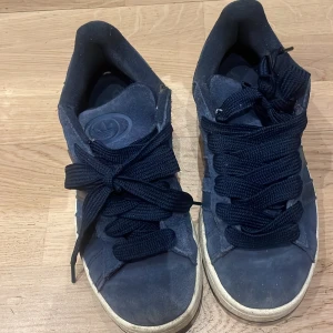 Blå adidas campus - Säljer ett par blå sneakers från Adidas med snörning. Skorna har en klassisk design med vit sula och är perfekta för vardagsbruk. De är tillverkade i mocka och har en bekväm passform. Jag köpte de höstas 2024 och förutom slitningarna läst uppe vid snöret som sysn på bilden så är de i jätte bra skick. De är i storlek 39,1/3 och passar även 38.