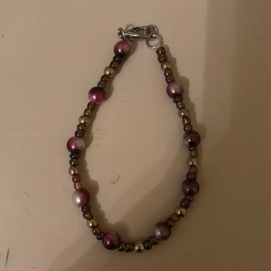 Färgglatt pärlarmband - Säljer ett färgglatt armband med pärlor i olika nyanser av lila, guld och metalfärgat. Armbandet har en praktisk karbinhake som gör det enkelt att ta på och av. Ej nickelfritt! 