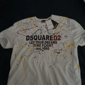 Vit t-shirt från Dsquared2 - Säljer en vit t-shirt från Dsquared2 med färgstänk i gult, rött och svart. Framsidan har texten 'LET YOUR DREAMS TAKE FLIGHT est. 1995' och baksidan har en stor logotyp. Perfekt för en cool och avslappnad stil.