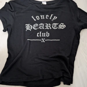 Svart t-shirt med tryck - Säljer en svart t-shirt med texten 'lonely HEARTS club' i vitt tryck. T-shirten har en avslappnad stil med korta ärmar och rund hals. Perfekt för en casual look.