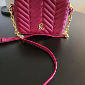 Vinröd axelväska från Ralph Lauren - Snygg vinröd axelväska från Ralph Lauren i skinn med quiltad design. Väskan har en elegant guldkedja och dragkedja upptill. Helt oanvänd 